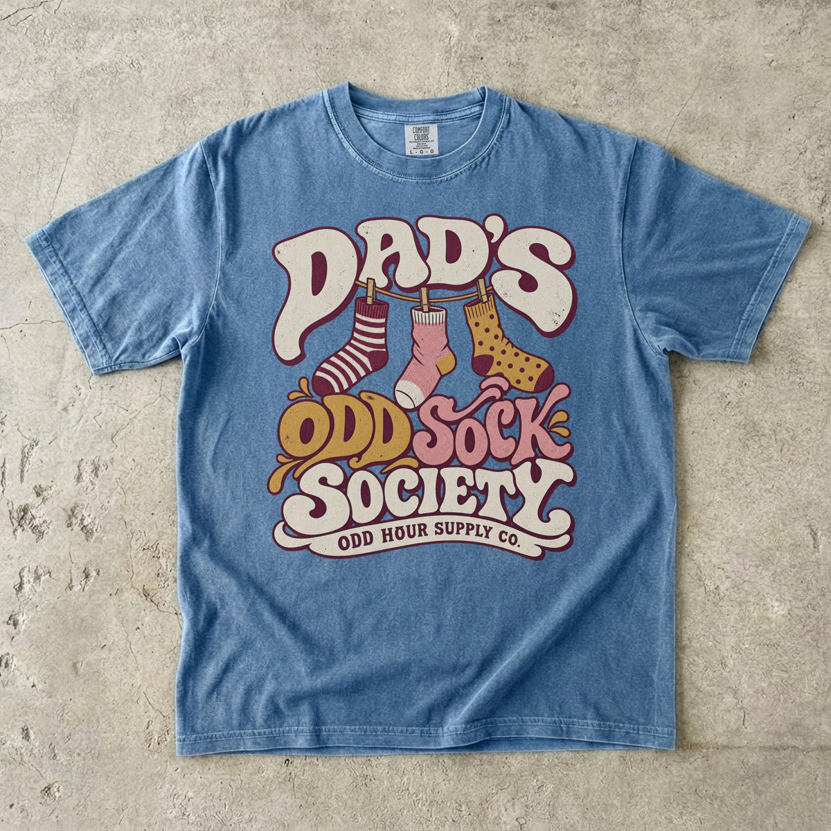 Blue Jean Comfort Colors 1717 T-shirt with three patterned socks on a clothesline and Dad’s Odd Sock Society lettering in cream, pink, and mustard yellow.