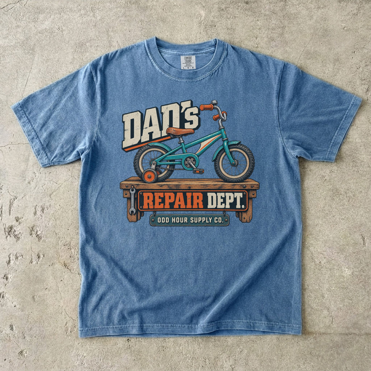 Blue Jean Comfort Colors 1717 T-shirt with a turquoise child’s bicycle with a training wheel on a workbench and Dad’s Repair Dept. lettering.