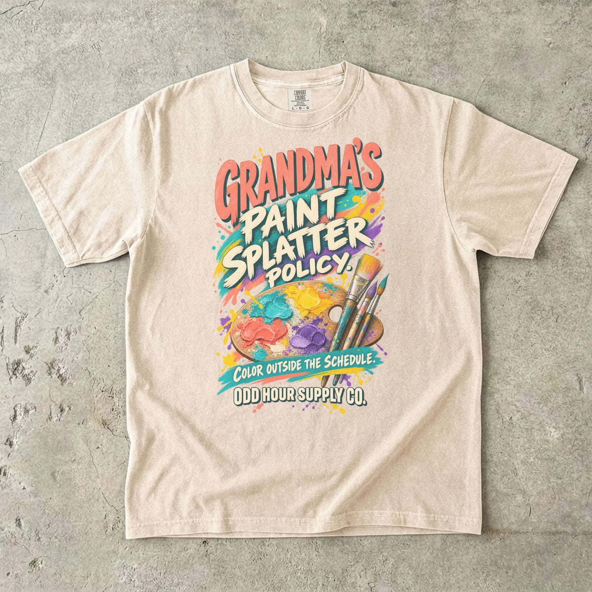 Ivory Comfort Colors 1717 T-shirt with an artist’s palette, paintbrushes, paint splashes, and Grandma’s Paint Splatter Policy and Color Outside the Schedule lettering.