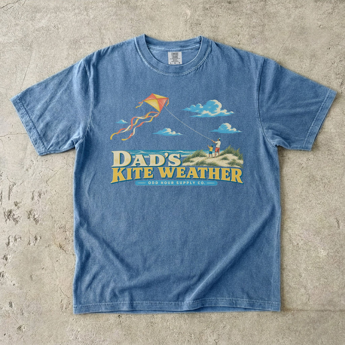 Blue Jean Comfort Colors 1717 T-shirt with a dad and child in beach dunes, an orange and yellow kite, sea, clouds, and Dad’s Kite Weather lettering.