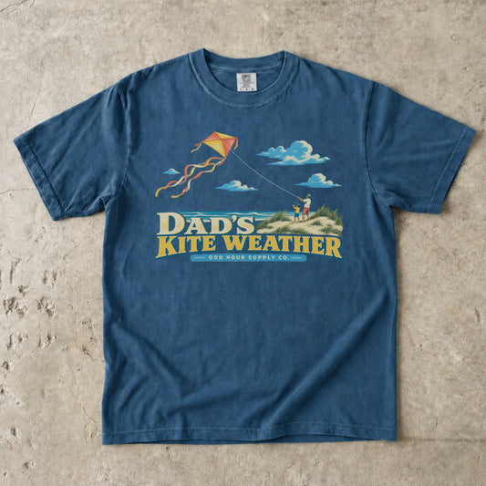 Midnight Comfort Colors 1717 T-shirt with a dad and child in beach dunes, an orange and yellow kite, sea, clouds, and Dad’s Kite Weather lettering.