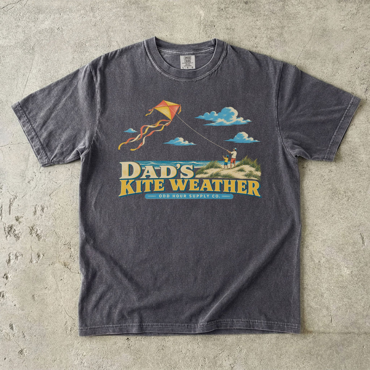 Pepper Comfort Colors 1717 T-shirt with a dad and child in beach dunes, an orange and yellow kite, sea, clouds, and Dad’s Kite Weather lettering.