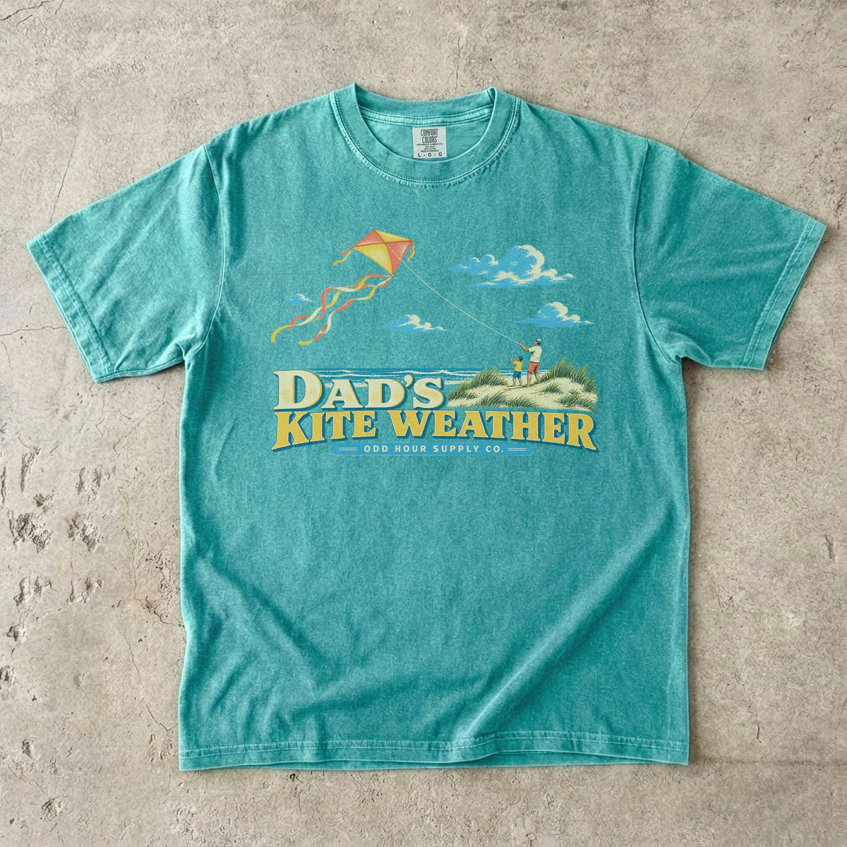 Seafoam Comfort Colors 1717 T-shirt with a dad and child in beach dunes, an orange and yellow kite, sea, clouds, and Dad’s Kite Weather lettering.