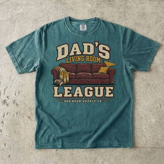 Blue Spruce Comfort Colors 1717 T-shirt with a reddish-brown couch, a football, a yellow blanket, and Dad’s Living Room League lettering.