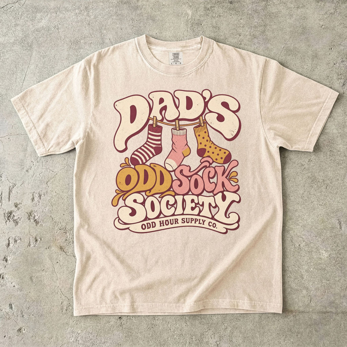 Ivory Comfort Colors 1717 T-shirt with three patterned socks on a clothesline and Dad’s Odd Sock Society lettering in cream, pink, and mustard yellow.