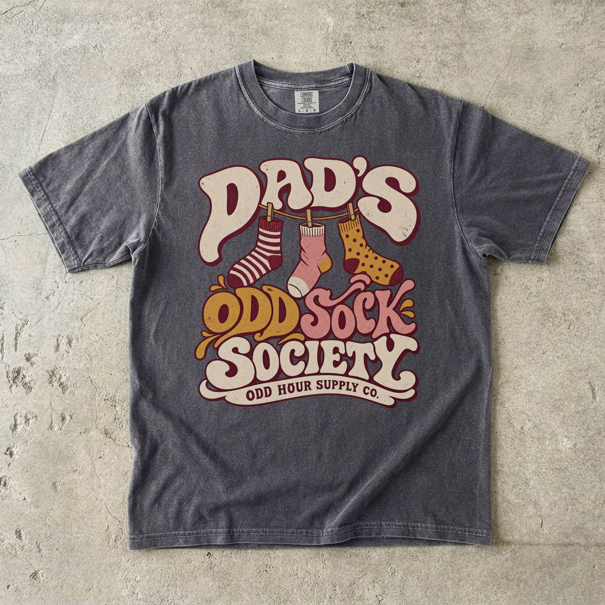 Pepper Comfort Colors 1717 T-shirt with three patterned socks on a clothesline and Dad’s Odd Sock Society lettering in cream, pink, and mustard yellow.
