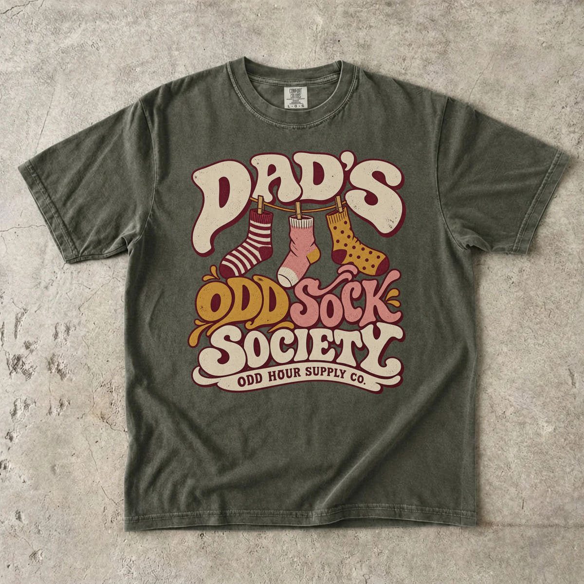 Sage Comfort Colors 1717 T-shirt with three patterned socks on a clothesline and Dad’s Odd Sock Society lettering in cream, pink, and mustard yellow.