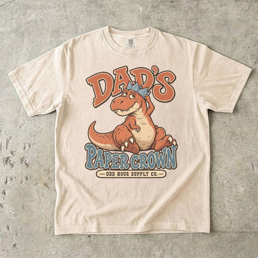 Ivory Comfort Colors 1717 T-shirt with a seated orange dinosaur, a blue paper crown, and Dad’s Paper Crown lettering.
