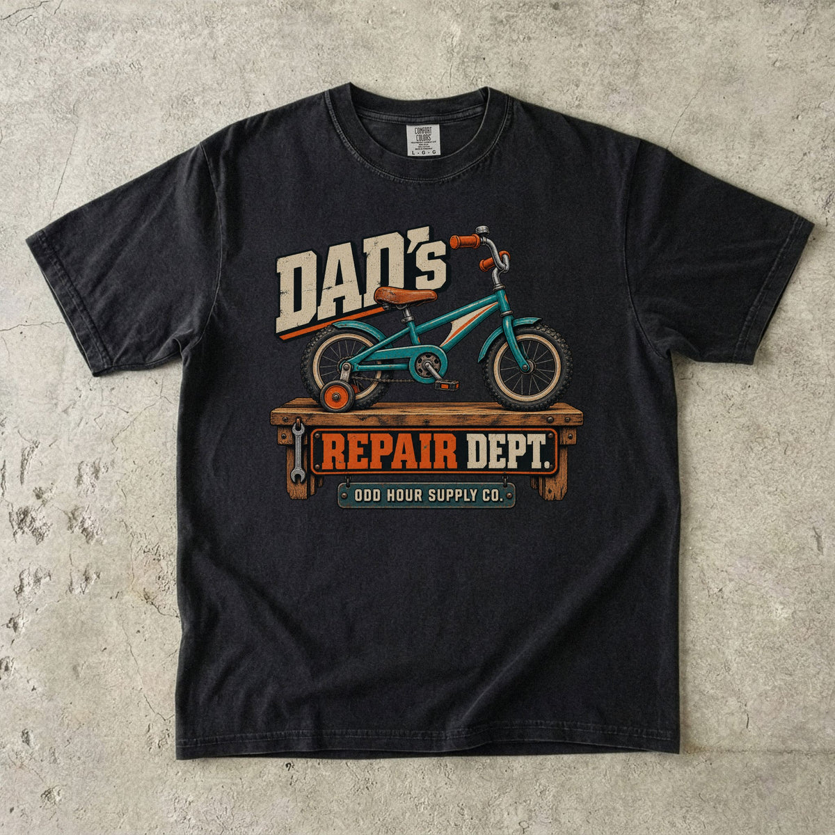 Black Comfort Colors 1717 T-shirt with a turquoise child’s bicycle with a training wheel on a workbench and Dad’s Repair Dept. lettering.