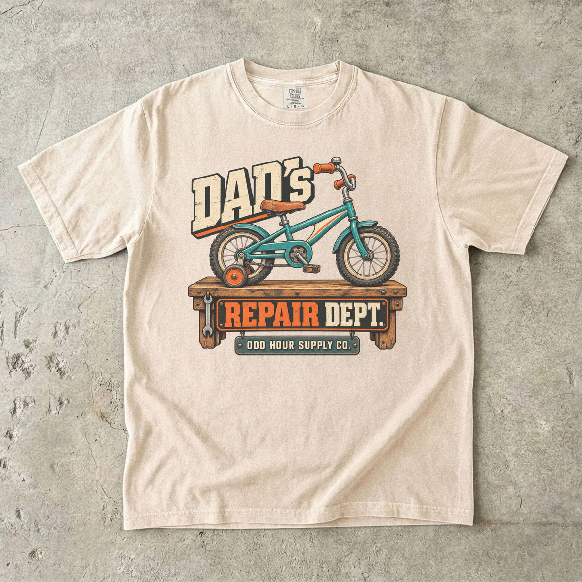 Ivory Comfort Colors 1717 T-shirt with a turquoise child’s bicycle with a training wheel on a workbench and Dad’s Repair Dept. lettering.