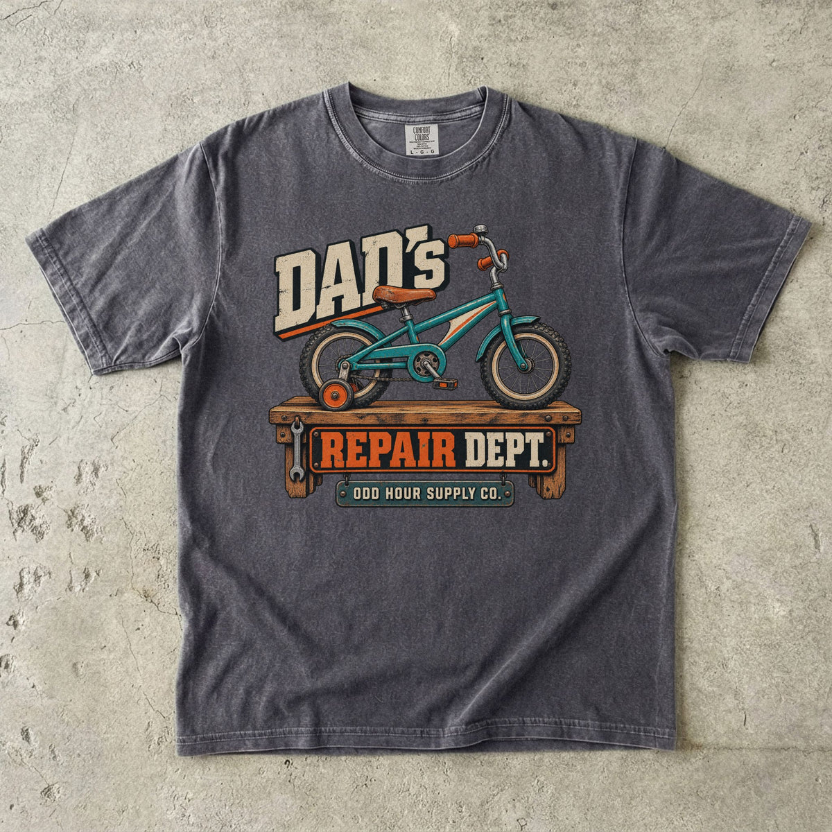 Pepper Comfort Colors 1717 T-shirt with a turquoise child’s bicycle with a training wheel on a workbench and Dad’s Repair Dept. lettering.