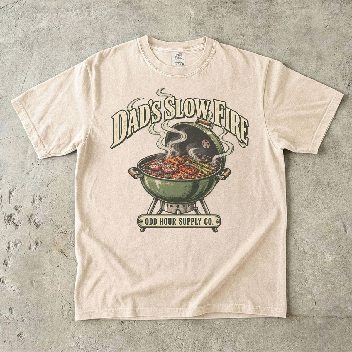 Ivory Comfort Colors 1717 T-shirt with a green kettle grill, vegetables on the grate, curling smoke, and Dad’s Slow Fire lettering.