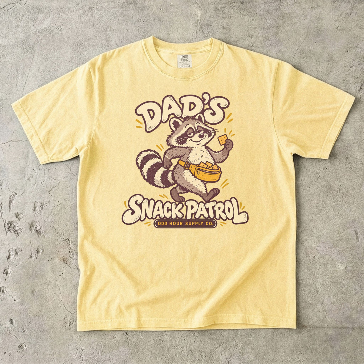Butter Comfort Colors 1717 T-shirt with a raccoon, a yellow waist pack full of crackers, and cream Dad’s Snack Patrol lettering.