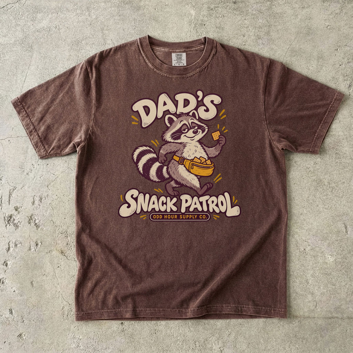 Espresso Comfort Colors 1717 T-shirt with a raccoon, a yellow waist pack full of crackers, and cream Dad’s Snack Patrol lettering.