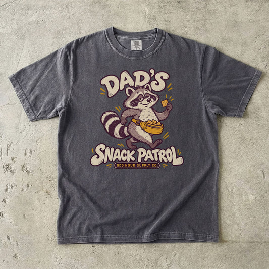 Pepper Comfort Colors 1717 T-shirt with a raccoon, a yellow waist pack full of crackers, and cream Dad’s Snack Patrol lettering.