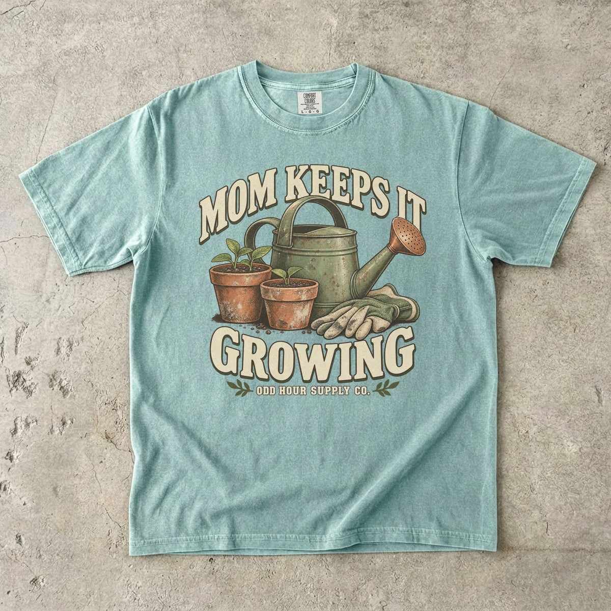 Bay Comfort Colors 1717 T-shirt with a green watering can, two terracotta pots with seedlings, gardening gloves, and Mom Keeps It Growing lettering.
