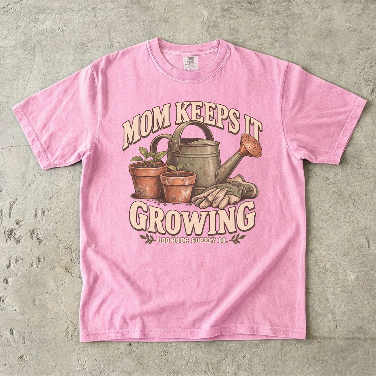 Blossom Comfort Colors 1717 T-shirt with a green watering can, two terracotta pots with seedlings, gardening gloves, and Mom Keeps It Growing lettering.
