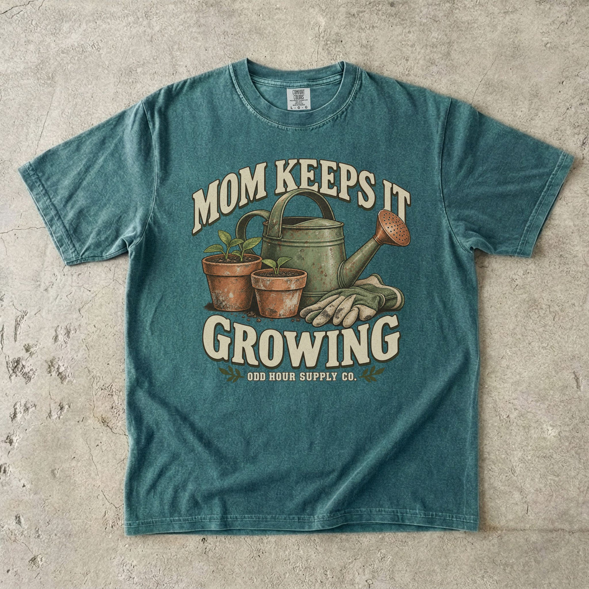 Blue Spruce Comfort Colors 1717 T-shirt with a green watering can, two terracotta pots with seedlings, gardening gloves, and Mom Keeps It Growing lettering.