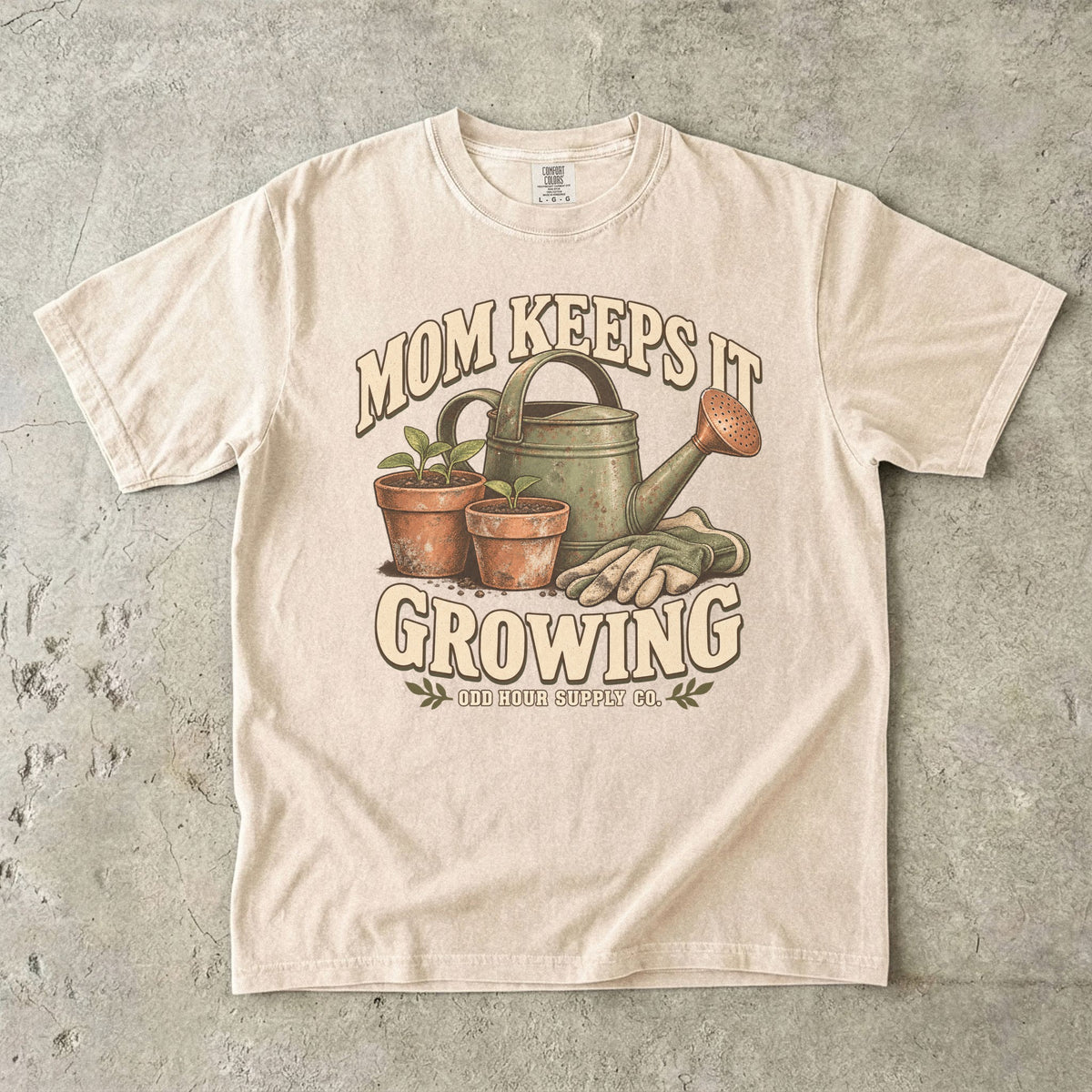 Ivory Comfort Colors 1717 T-shirt with a green watering can, two terracotta pots with seedlings, gardening gloves, and Mom Keeps It Growing lettering.