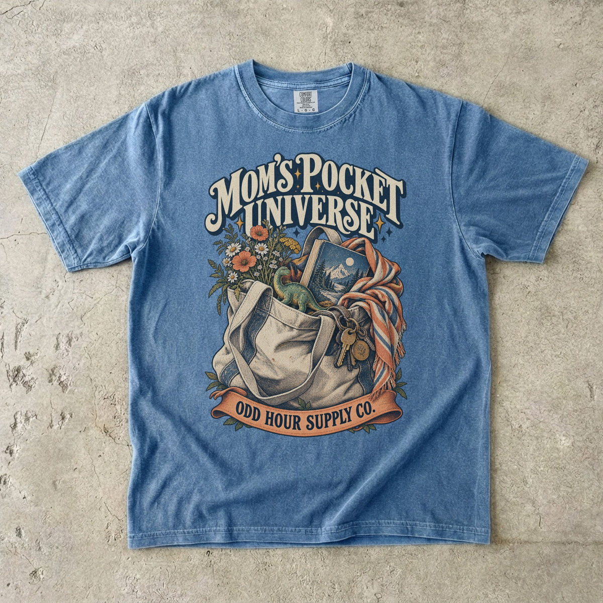 Blue Jean Comfort Colors 1717 T-shirt with a tote bag holding flowers, a book, a toy dinosaur, and keys beneath Mom’s Pocket Universe lettering.