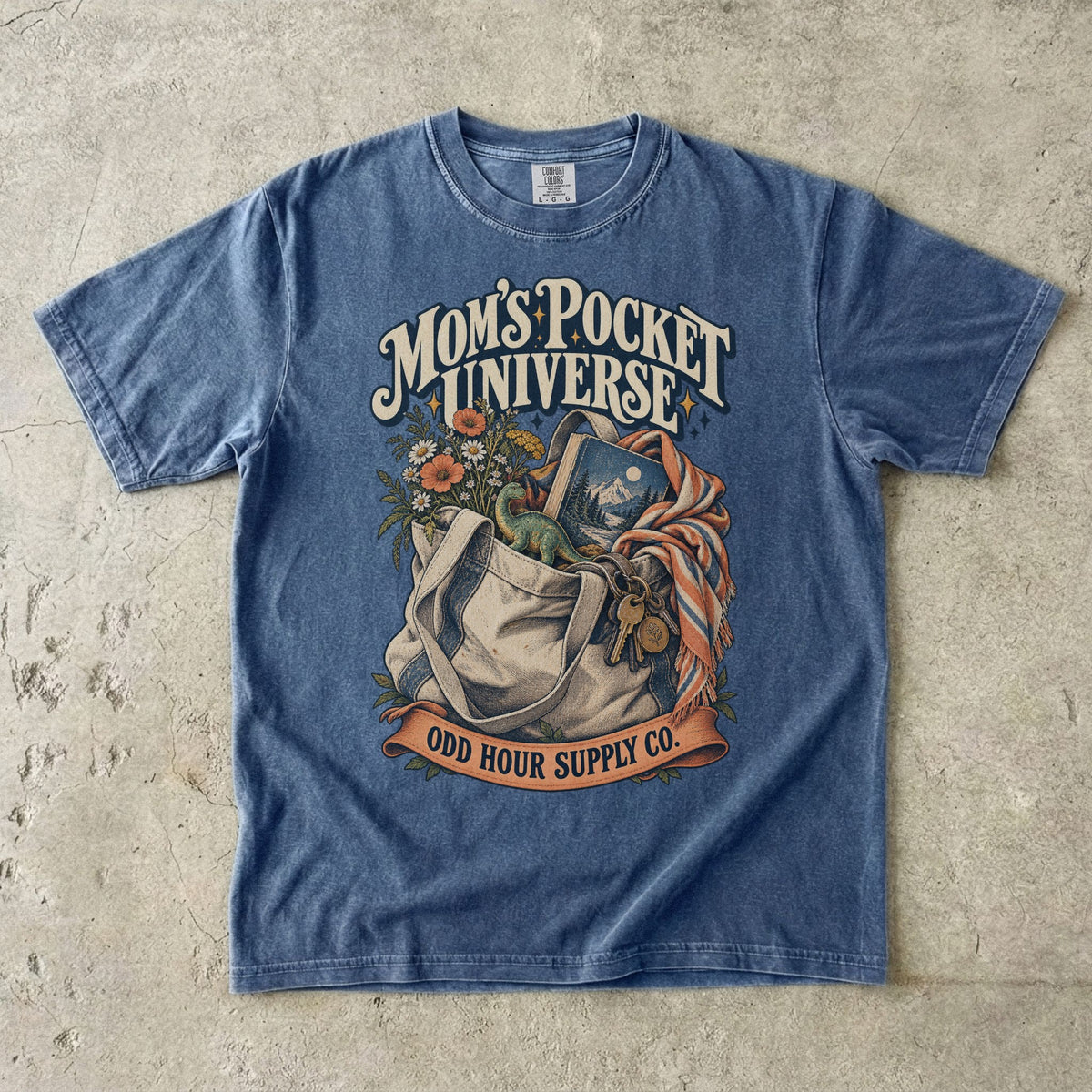 Denim Comfort Colors 1717 T-shirt with a tote bag holding flowers, a book, a toy dinosaur, and keys beneath Mom’s Pocket Universe lettering.