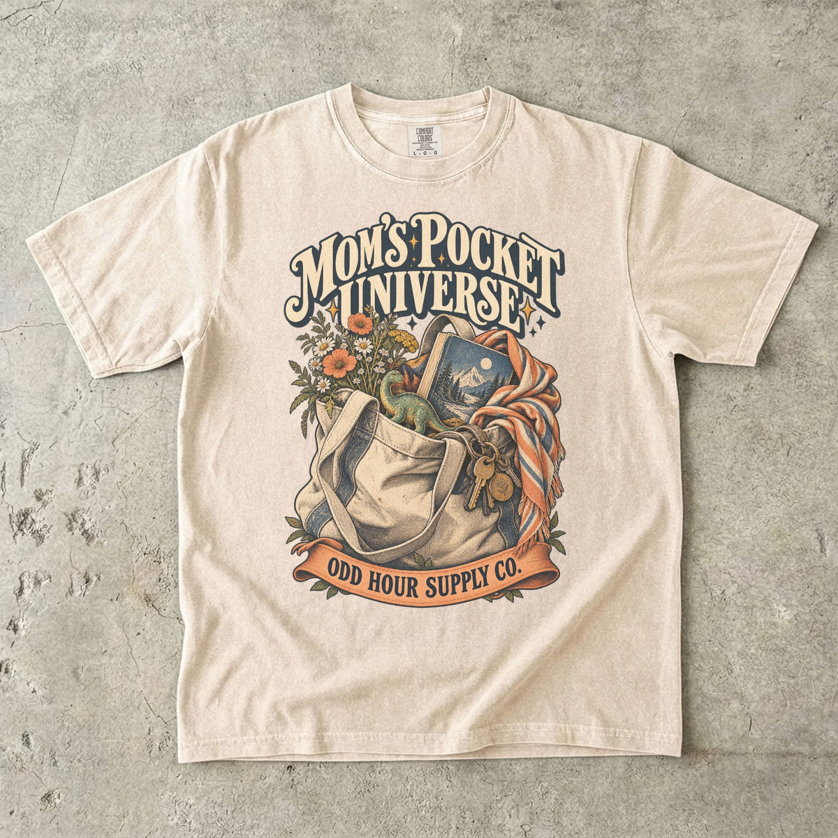 Ivory Comfort Colors 1717 T-shirt with a tote bag holding flowers, a book, a toy dinosaur, and keys beneath Mom’s Pocket Universe lettering.