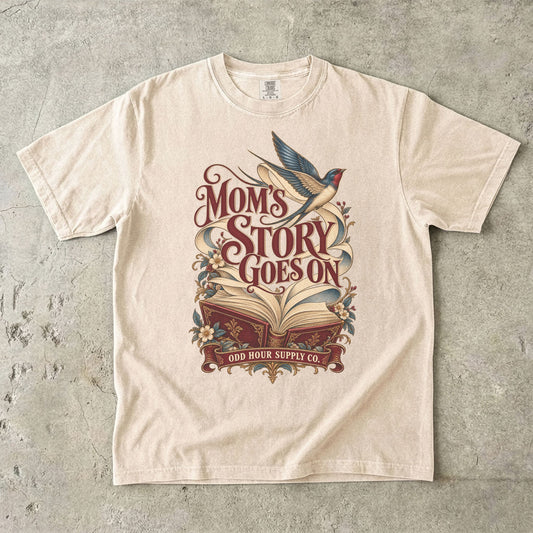 Ivory Comfort Colors 1717 T-shirt with an open red book, flowing pages, a flying swallow, flowers, and Mom’s Story Goes On lettering.