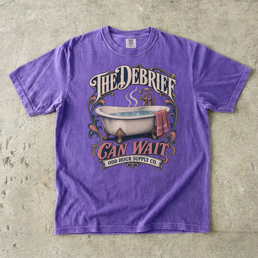 Violet T-shirt with a vintage bathtub, THE DEBRIEF CAN WAIT lettering and decorative Odd Hour Supply Co. branding.