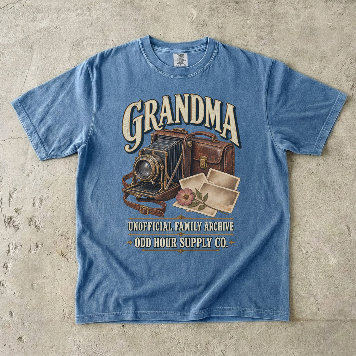 Blue Jean Comfort Colors 1717 T-shirt with a vintage folding camera, brown camera case, photo prints, a flower, and Grandma Unofficial Family Archive lettering.