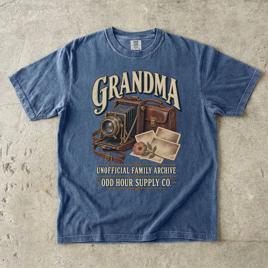 Denim Comfort Colors 1717 T-shirt with a vintage folding camera, brown camera case, photo prints, a flower, and Grandma Unofficial Family Archive lettering.