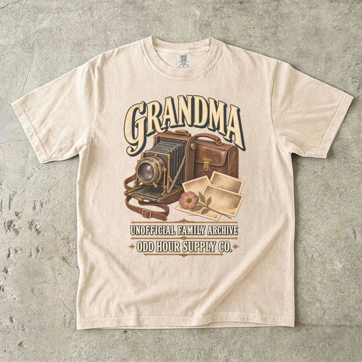 Ivory Comfort Colors 1717 T-shirt with a vintage folding camera, brown camera case, photo prints, a flower, and Grandma Unofficial Family Archive lettering.